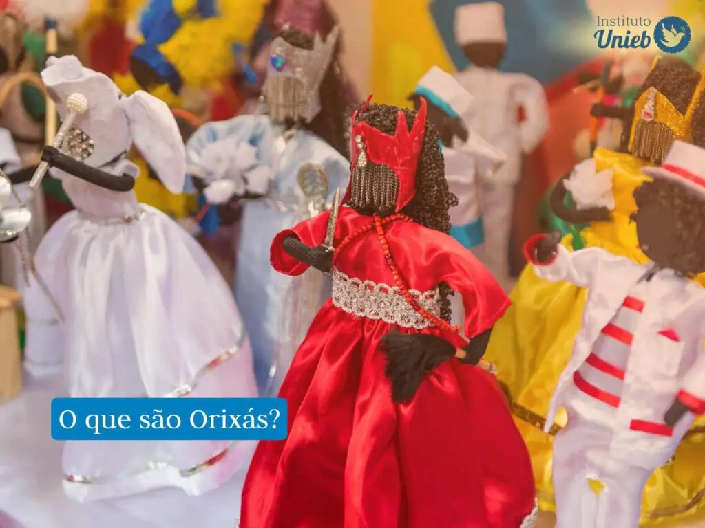 bonecos ou representações em miniatura de Entidades cultuadas nas religiões afro-brasileiras, na imagem vemos Orixás como Iemanjá, Iansã, Nanã, Oxum e outras Entidades Espirituais, há uma frase com fundo azul que diz "o que são Orixás?" e um logo acima do Instituto Unieb