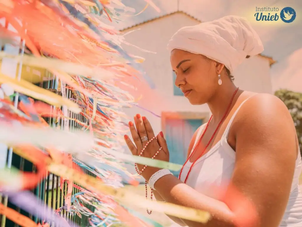 mulher com mãos fechadas em prece com roupa branca e pano branco na cabeça, praticante da Umbanda, de frente para grade com fitas de cetim amarradas