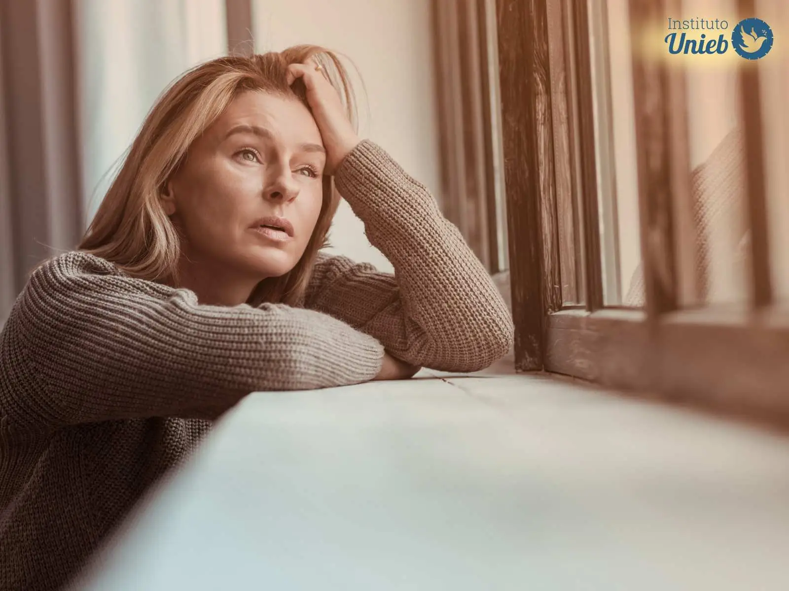 Mulher vestindo suéter com os braços apoiados em uma base e uma mão na cabeça, enquanto olha pela janela com expressão de tristeza