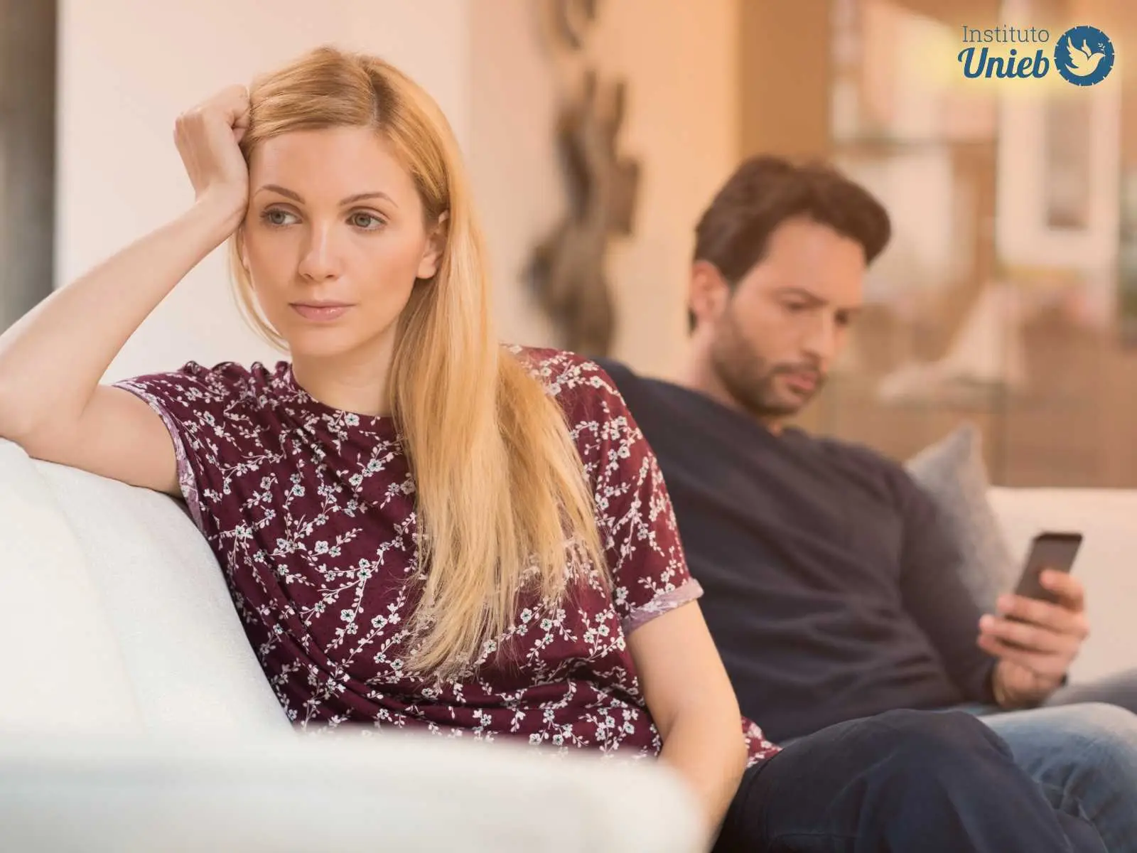 Mulher com a mão na cabeça de costas para homem ao fundo mexendo no celular