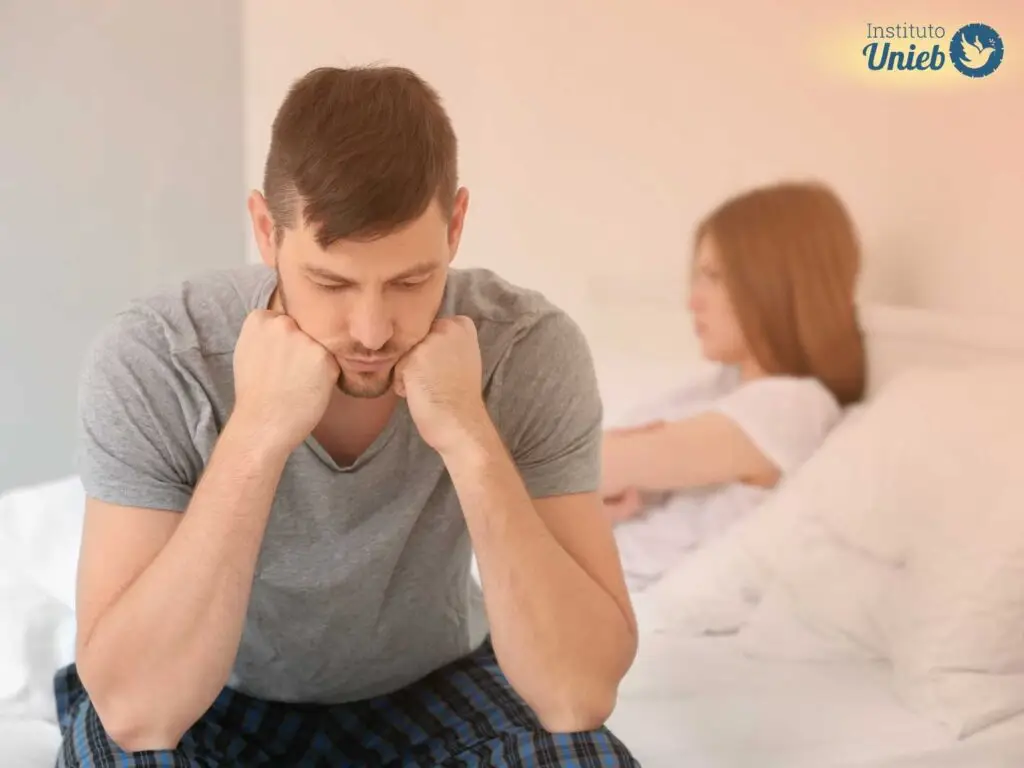 Homem com a cabeça apoiada nos braços pensativo e mulher ao fundo com os braços cruzados