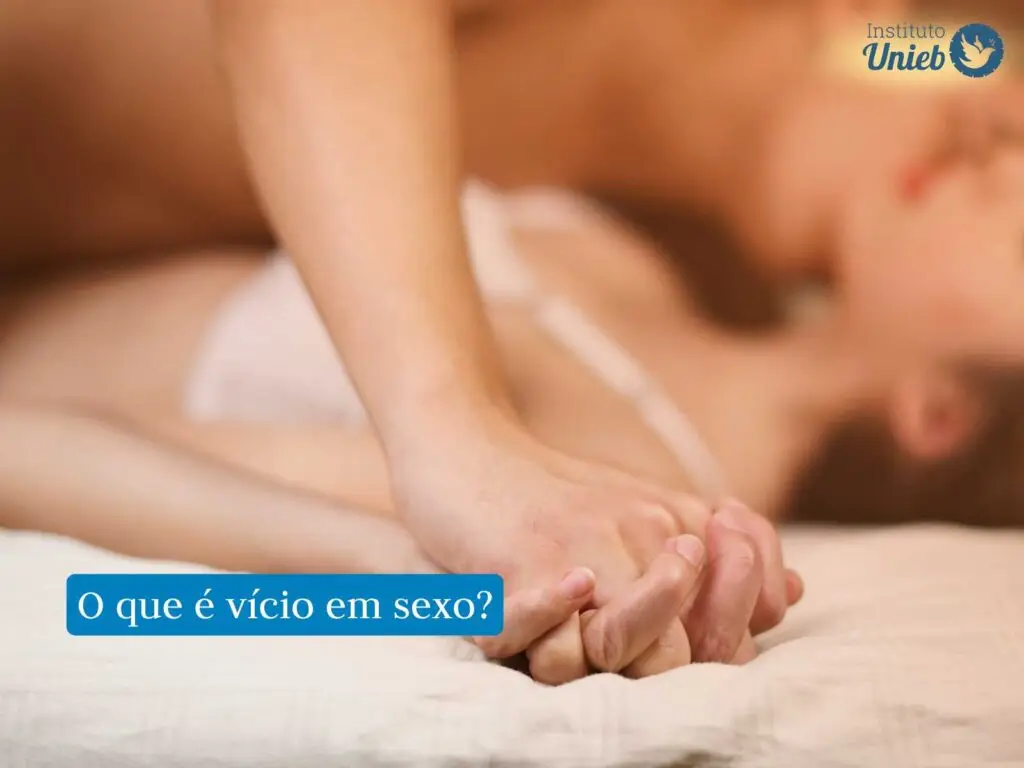 Mãos de um casal entrelaçadas e apoiadas na cama e ao fundo o casal em momento de intimidade