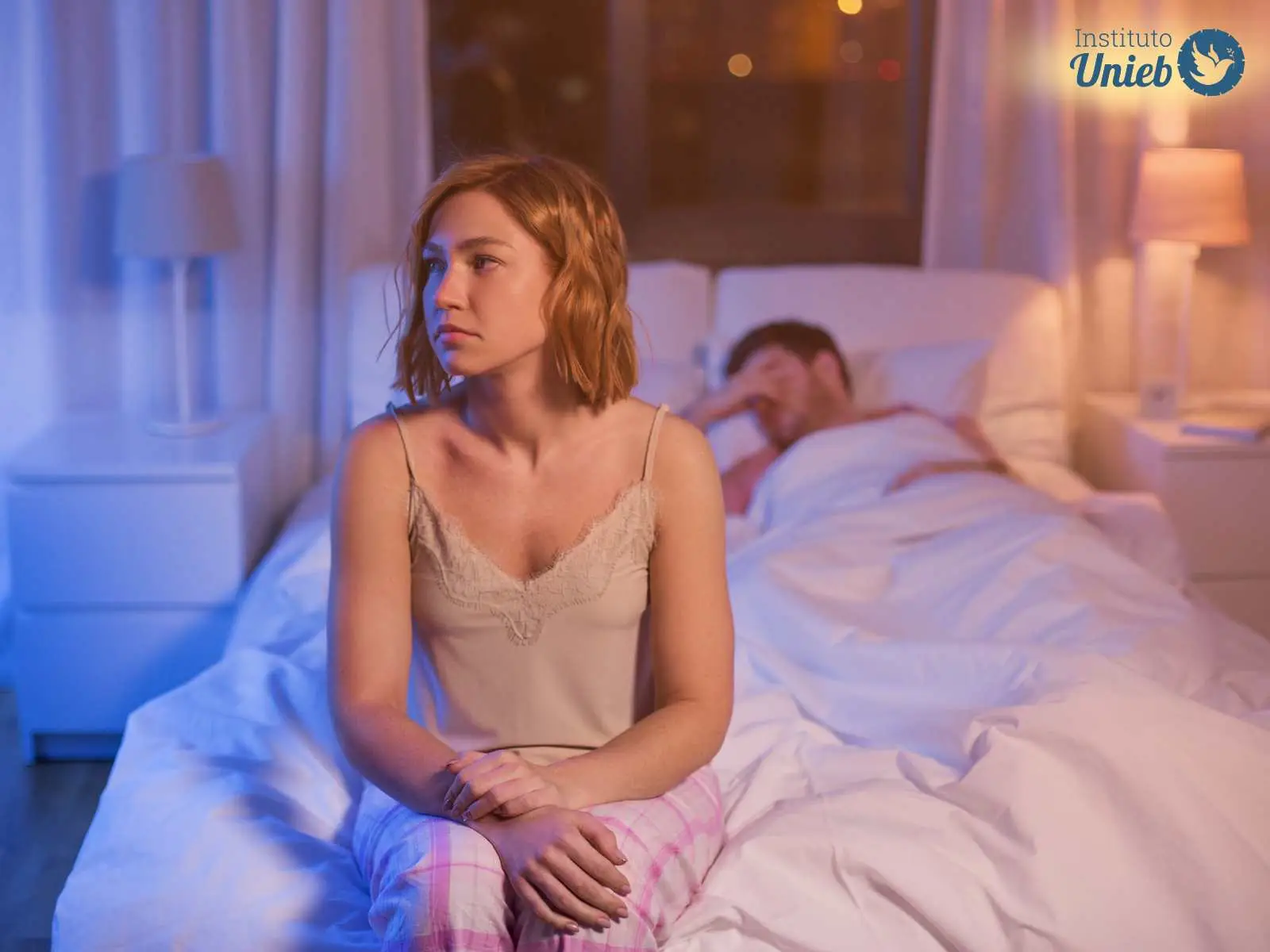 Mulher sentada na cama com expressão pensativa com homem deitado na cama em baixo de edredom branco ao fundo