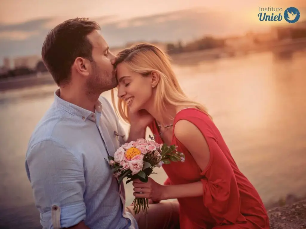 Homem de cabelo preto e camisa azul claro beijando a testa de uma mulher loira com vestido vermelho segurando um buquê de flores coloridas ao ar livre