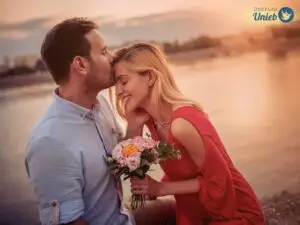 Homem de cabelo preto e camisa azul claro beijando a testa de uma mulher loira com vestido vermelho segurando um buquê de flores coloridas ao ar livre