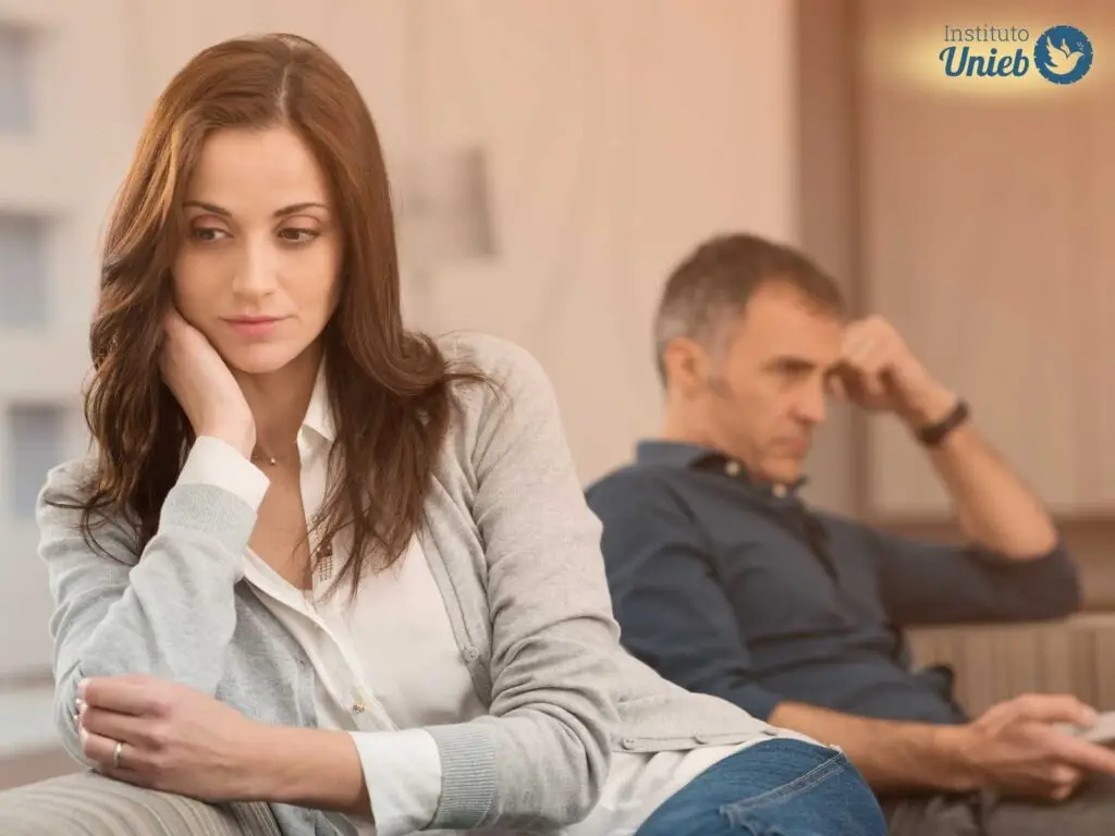 Mulher sentada em sofá com camisa branca, braço apoiado no rosto com expressão triste e homem ao funco com cabeça apoiada no braço