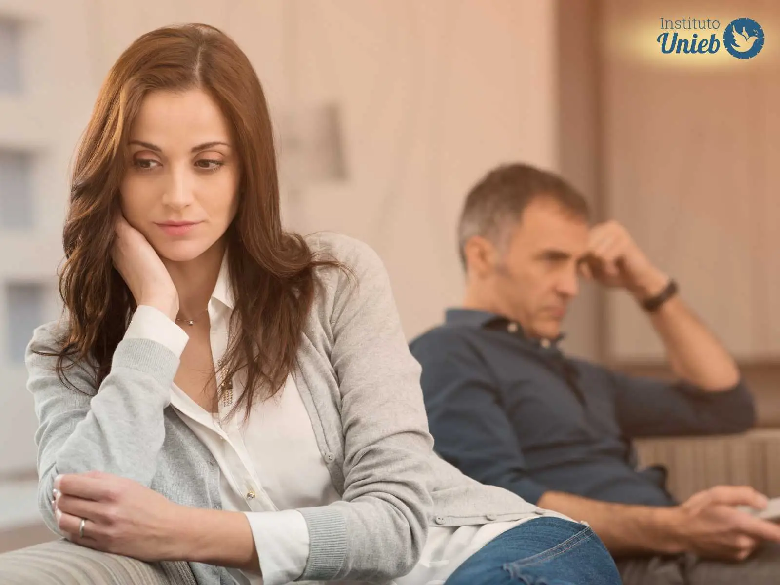 Blog Mulher sentada em sofá com camisa branca, braço apoiado no rosto com expressão triste e homem ao funco com cabeça apoiada no braço