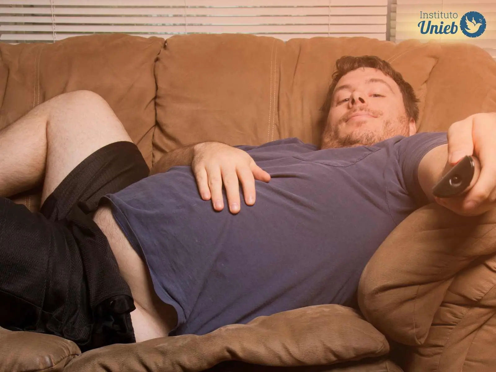 Homem vestindo camiseta azul e shorts preto deitado em sofá marrom