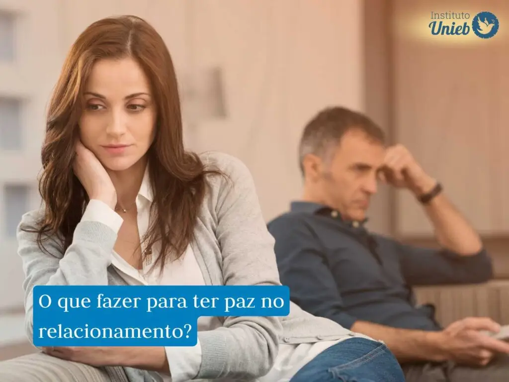 Mulher sentada em sofá com camisa branca, braço apoiado no rosto com expressão triste e homem ao funco com cabeça apoiada no braço