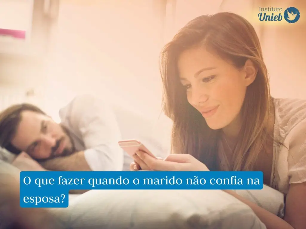 Mulher com celular na mão e sorriso no rosto deitada na cama com marido ao fundo deitado na cama e a observando