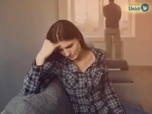 Mulher com a cabeça apoiada no braço e expressão triste com homem ao fundo de costas olhando pela janela