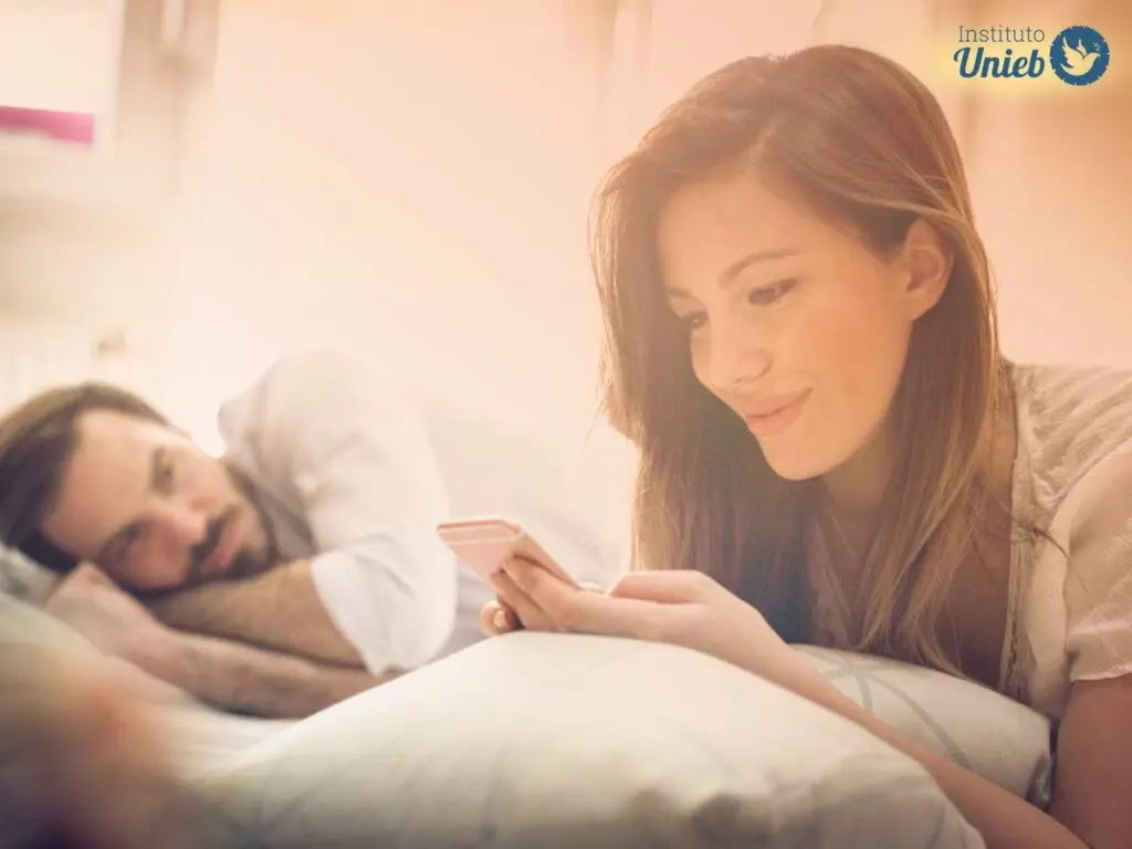 Mulher com celular na mão e sorriso no rosto deitada na cama com marido ao fundo deitado na cama e a observando