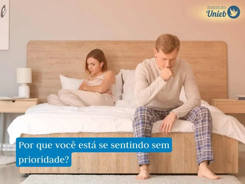 Mulher deitada na cama ao fundo e homem sentado ao pé da cama com a mão na boca e cabeça inclinada para baixo