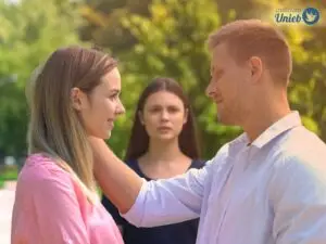 Casal olhando de frente para o outro em um ambiente aberto com mulher ao fundo olhando os dois com inveja
