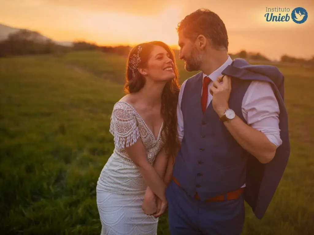 Homem e mulher vestidos de noivos em pose olhando um para o outro em um campo com sol se pondo ao fundo