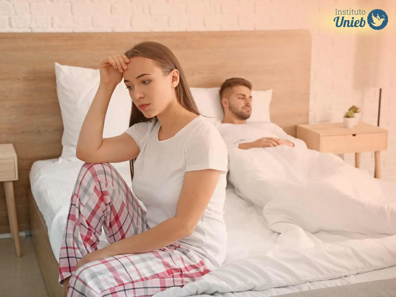 Homem deitado na cama embaixo das cobertas e mulher sentada ao pé da cama com cabeça apoiada na mão direita e joelho