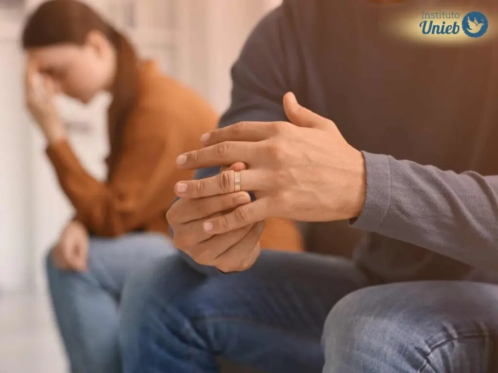 Homem sentado retirando aliança de casamento do dedo e mulher ao fundo desfocada com a mão na cabeça