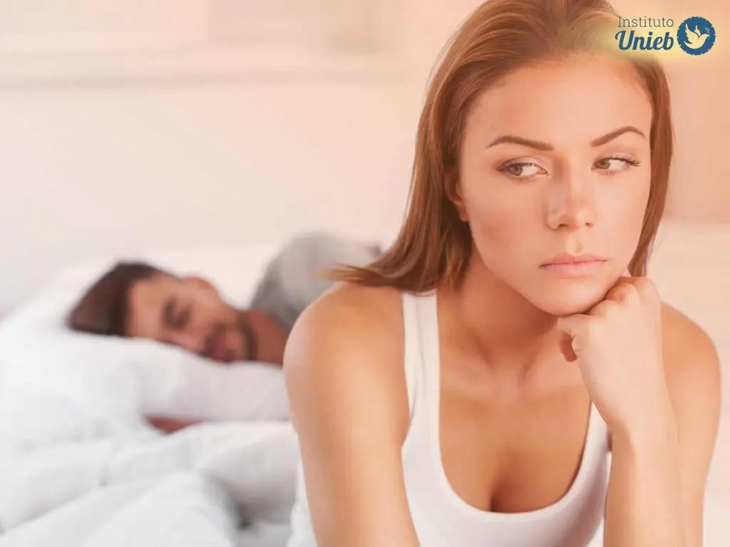 Mulher sentada na cama com a cabeça apoiada na mão e homem deitado na cama ao fundo dormindo