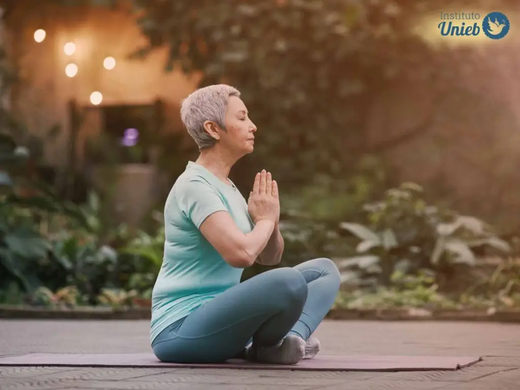 Mulher de meia idade sentada ao chão em posição de ioga com as mãos em prece vestindo camiseta verde claro e calça cinza de ginástica ao ar livre