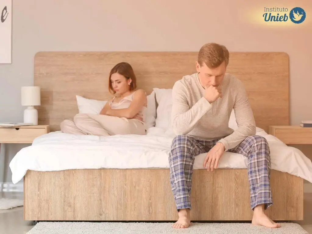 Mulher deitada na cama ao fundo e homem sentado ao pé da cama com a mão na boca e cabeça inclinada para baixo