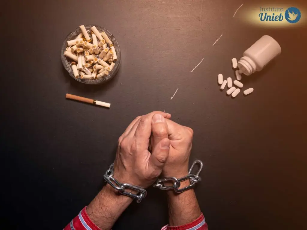 Mãos de um homem acorrentadas em cima da mesa ao lado de um cinzeiro, cigarro e alguns comprimidos - Instituto Unieb (Pai de santo Roberson Dariel) - Contato 11 96982-1121 - CEP 14801-040 - Av. Dom Pedro II 562, Centro, Araraquara, SP