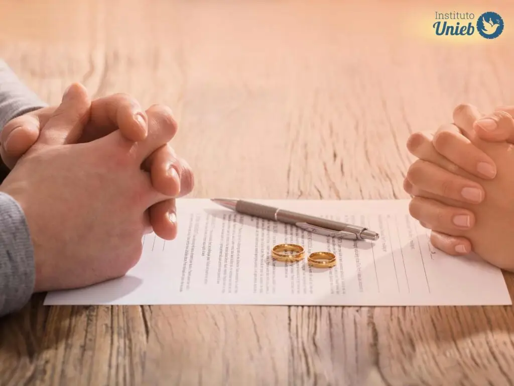 Homem e mulher com mãos fechadas em cima de uma mesa de madeira com papel de divórcio, alianças e caneta em cima