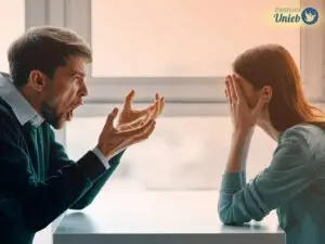 Mulher sentada de um lado da mesa com as mãos no rosto e braços apoiados na mesa e homem do outro lado com braços apoiados na mesa e gesticulando enquanto grita algo para a mulher