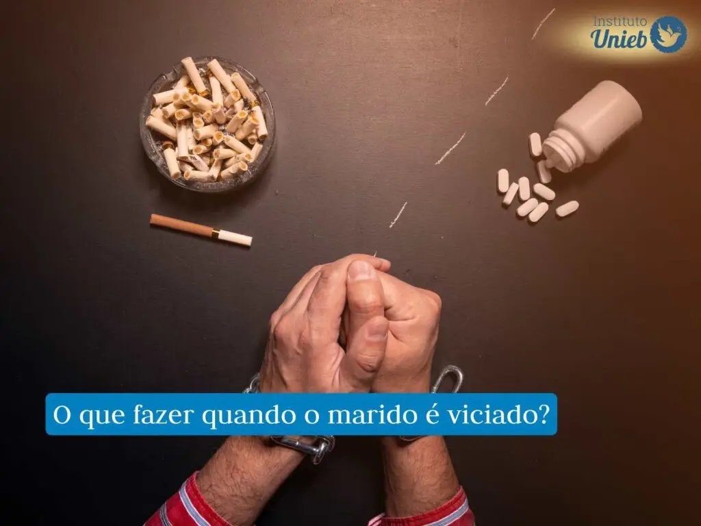 Mãos de um homem acorrentadas em cima da mesa ao lado de um cinzeiro, cigarro e alguns comprimidos