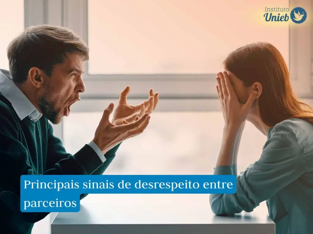 Mulher sentada de um lado da mesa com as mãos no rosto e braços apoiados na mesa e homem do outro lado com braços apoiados na mesa e gesticulando enquanto grita algo para a mulher