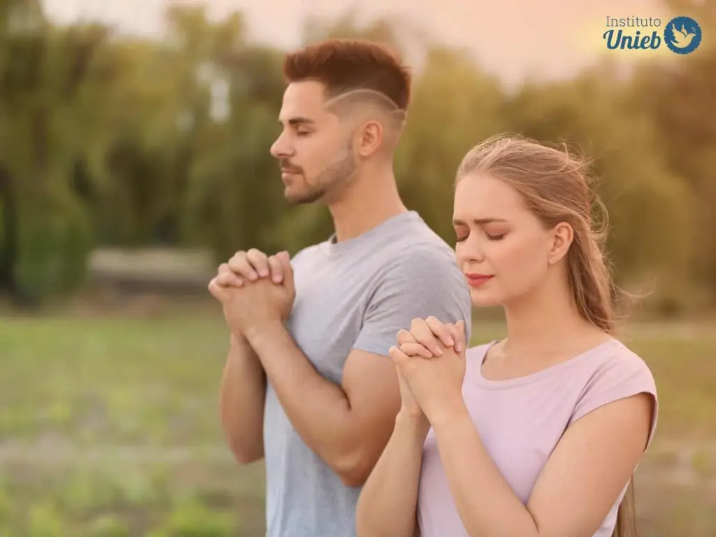 Homem e mulher em ambiente externo com mãos em prece e olhos fechados enquanto rezam