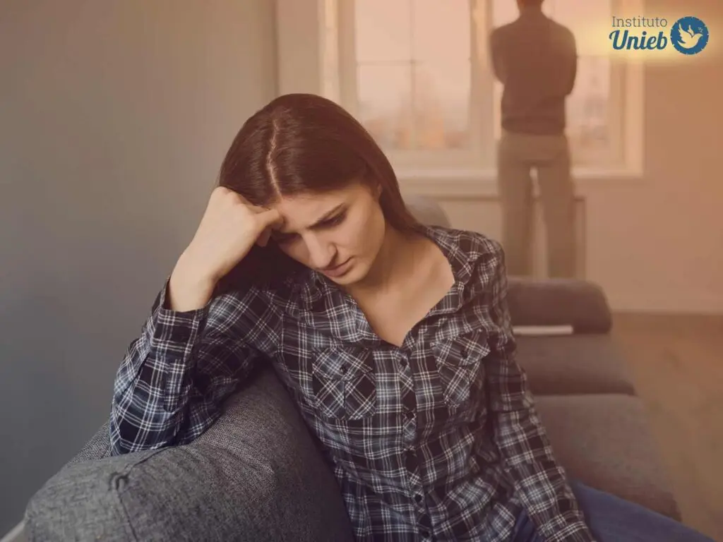 Mulher sentada em sofá com a cabeça apoiada na mão direita e expressão triste enquanto home olha para fora da janela ao fundo de costas para a mulher