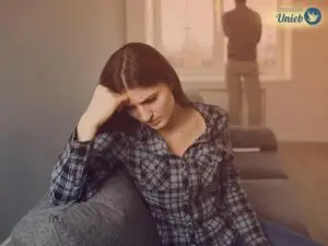 Mulher sentada em sofá com a cabeça apoiada na mão direita e expressão triste enquanto home olha para fora da janela ao fundo de costas para a mulher