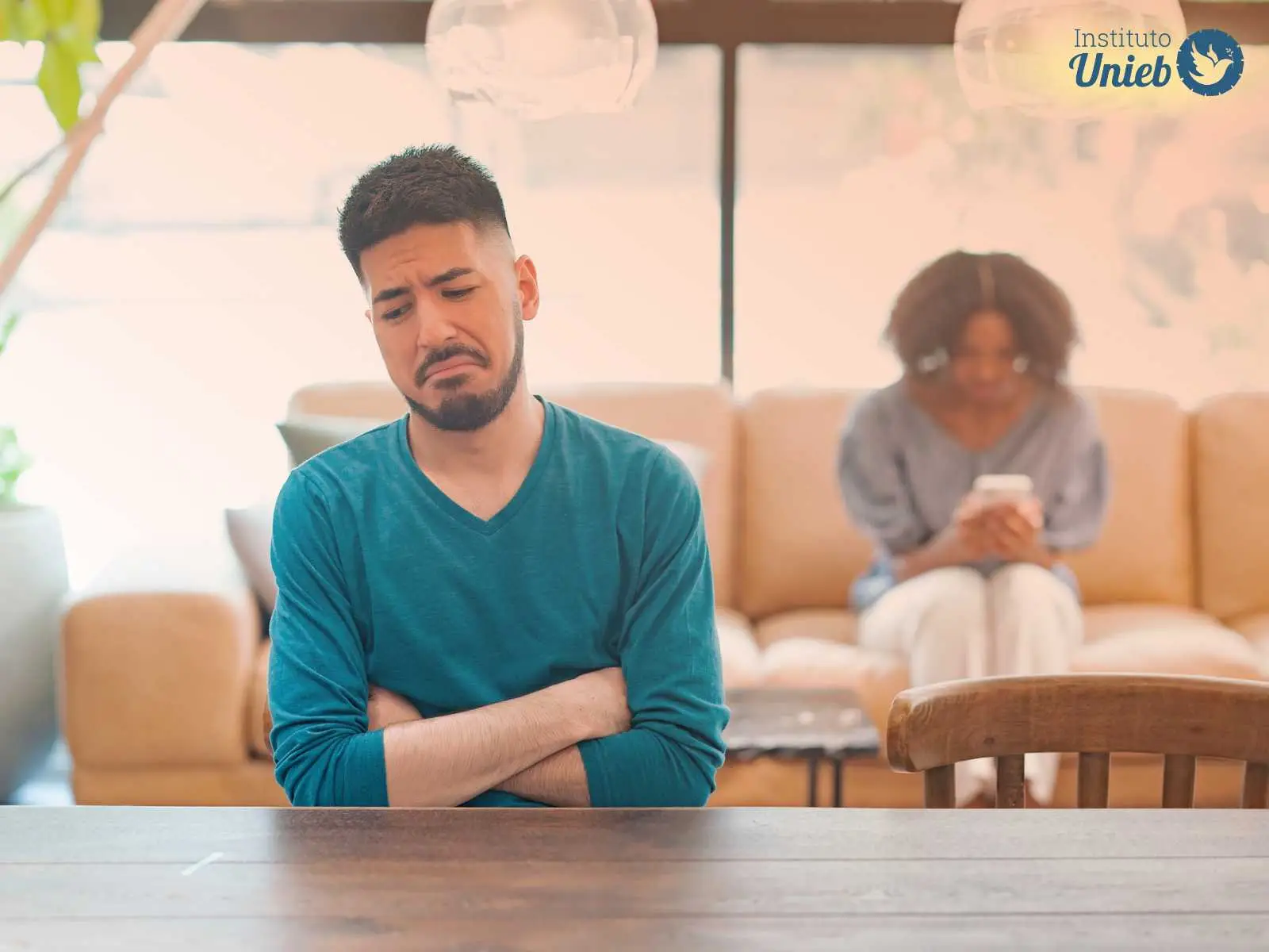 Homem sentado à mesa de braços cruzados e expressão de tristeza com mulher ao fundo sentada no sofá mexendo no celular