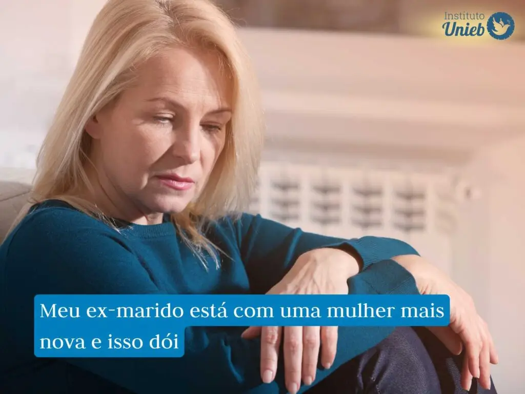 Mulher madura com cabelo loiro sentada com braços apoiados nas pernas vestindo blusa azul