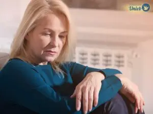 Mulher madura com cabelo loiro sentada com braços apoiados nas pernas vestindo blusa azul