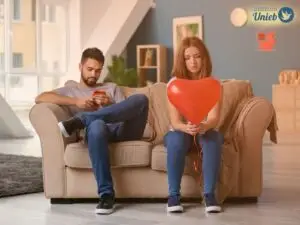 Homem e mulher sentados em sofá bege na sala enquanto homem mexe no celular e mulher segura um balão em formato de coração vermelho