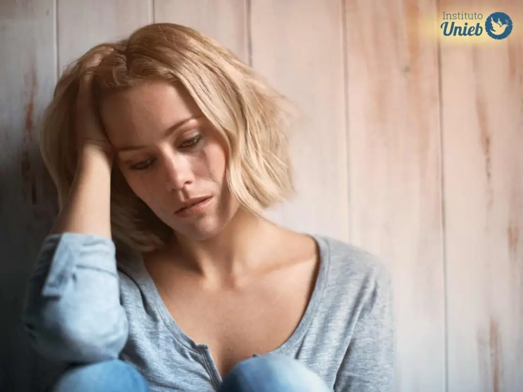 Mulher de blusa e calça azul sentada e encostada em parede de madeira com cabeça apoiada no braço direito