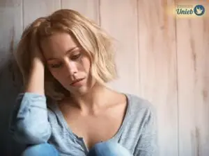 Mulher de blusa e calça azul sentada e encostada em parede de madeira com cabeça apoiada no braço direito