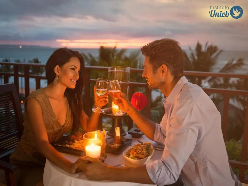 Homem e mulher sentados a mesa em jantar a luz de velas com taças nas mãos brindando e pôr do sol ao fundo