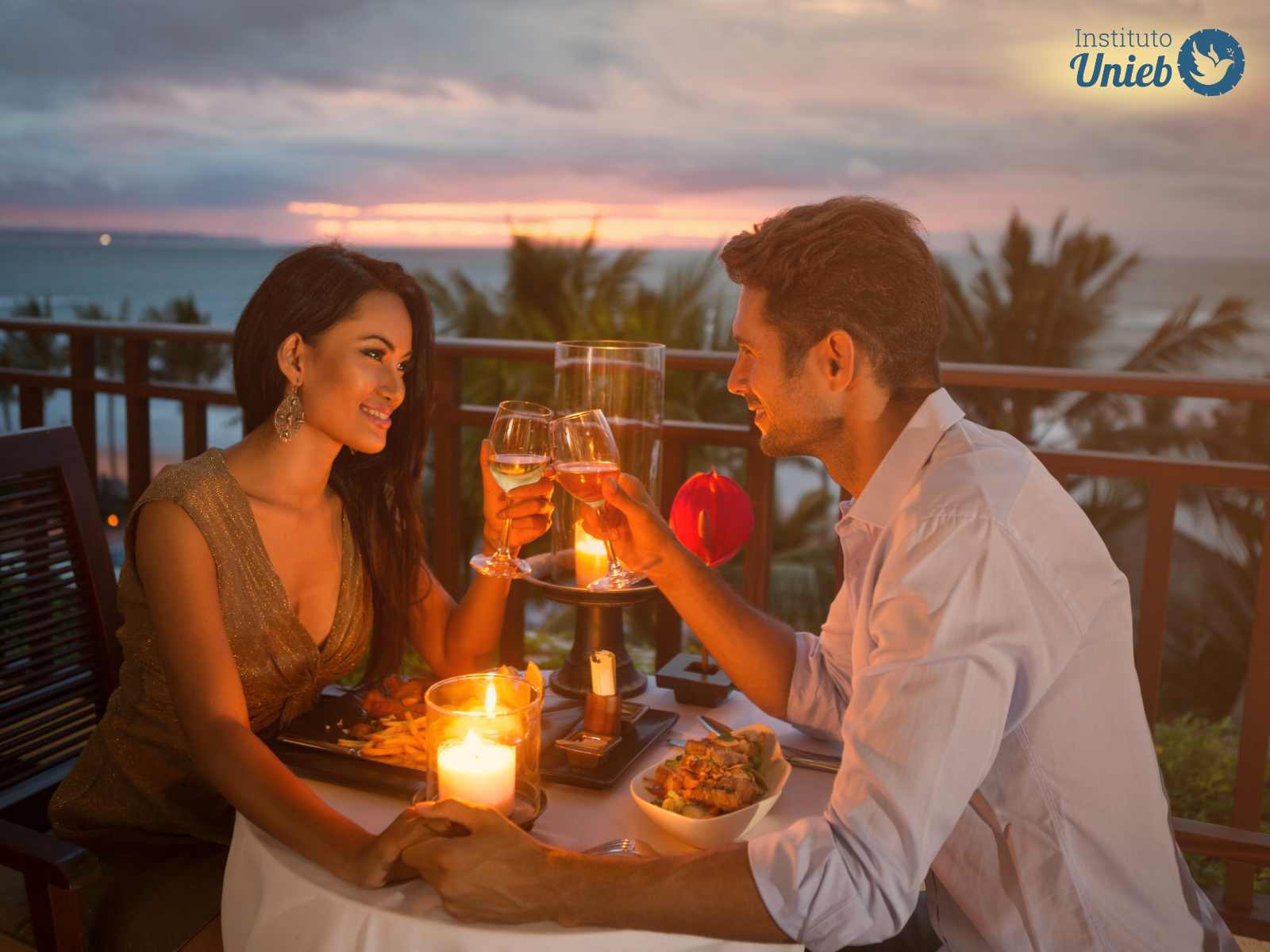 Homem e mulher sentados a mesa em jantar a luz de velas com taças nas mãos brindando e pôr do sol ao fundo
