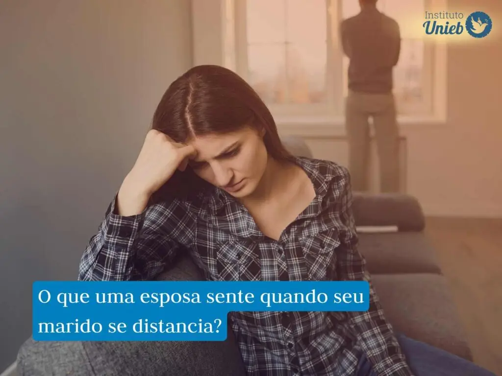 Mulher sentada em sofá com a cabeça apoiada na mão direita e expressão triste enquanto home olha para fora da janela ao fundo de costas para a mulher