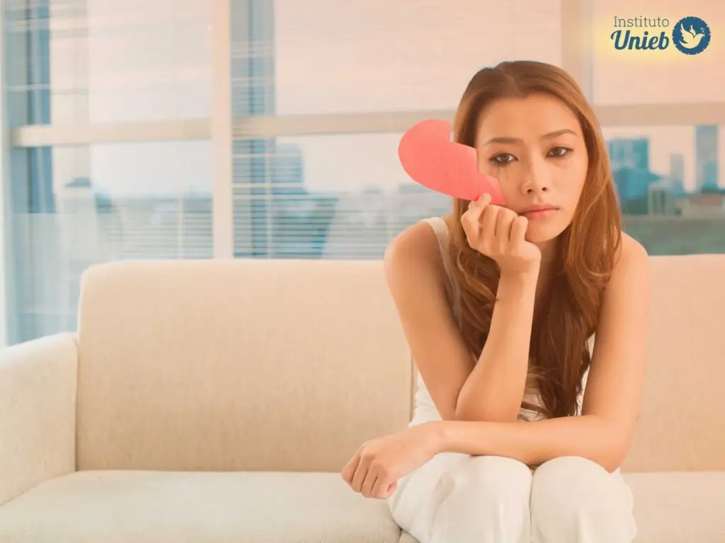 Mulher sentada em sofá com metade de um coração de papel na mão e com o rosto borrado de maquiagem após chorar