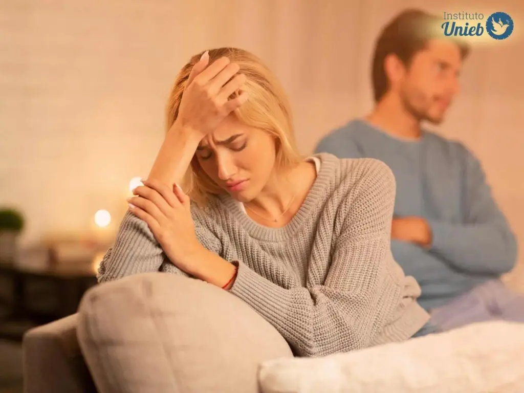 Casal de homem e mulher sentados no sofá, onde homem veste camiseta azul e está de braços cruzados e mulher com blusa cinza está com a cabeça apoiada na mão
