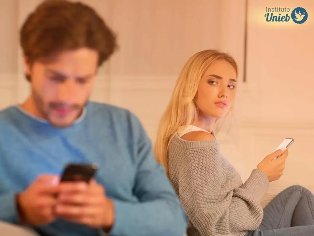 Homem sentado no sofá olhando para celular e mulher ao fundo com celular na mão olhando para seu marido desconfiada e triste
