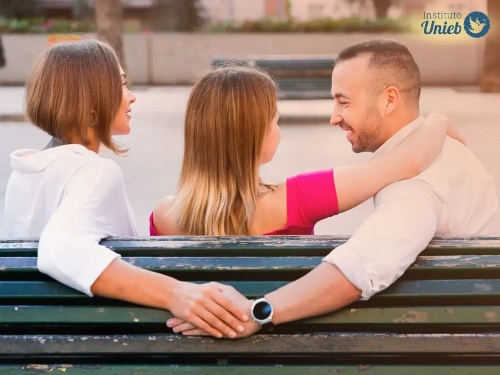 Casal de homem e mulher sentados em banco de uma praça onde a mulher está com braço nos ombros do homem e homem está tocando a mão de outra mulher sentada à esquerda