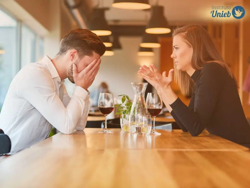 Homem e mulher sentados com braços apoiados em mesa de madeira com taças de vinho e com gestos de discussão