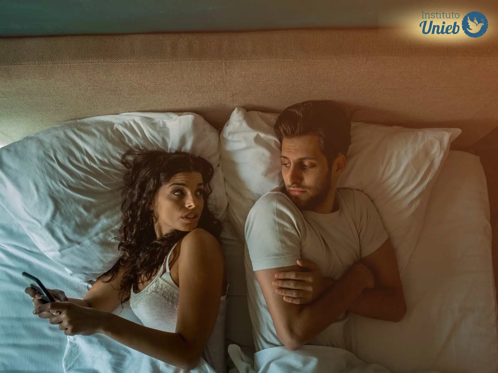 Casal deitado na cama, onde mulher mexe no celular e seu parceiro olha com desconfiança