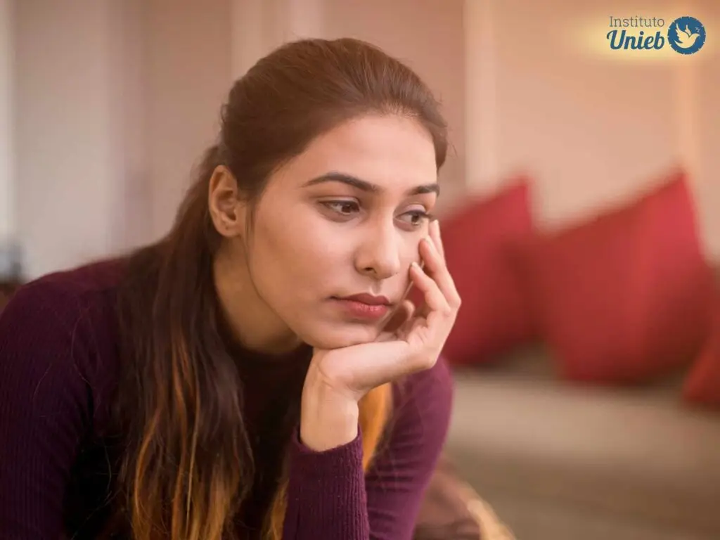 Mulher vestindo camiseta de manga comprida na cor vinho com a cabeça apoiada na mão esquerda com expressão de tristeza