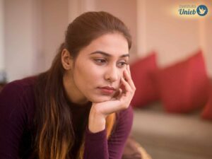 Mulher vestindo camiseta de manga comprida na cor vinho com a cabeça apoiada na mão esquerda com expressão de tristeza
