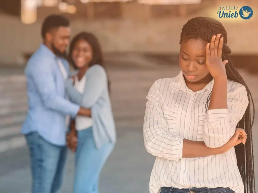 Mulher com mão no rosto e outro braço na cintura e ao fundo um casal sorrindo